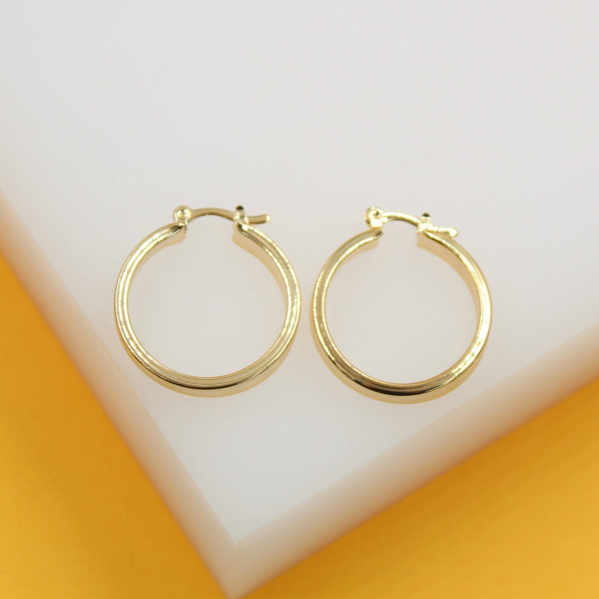 A pair of shiny gold hoop earrings rests neatly on a clean white display block against a vibrant yellow background.