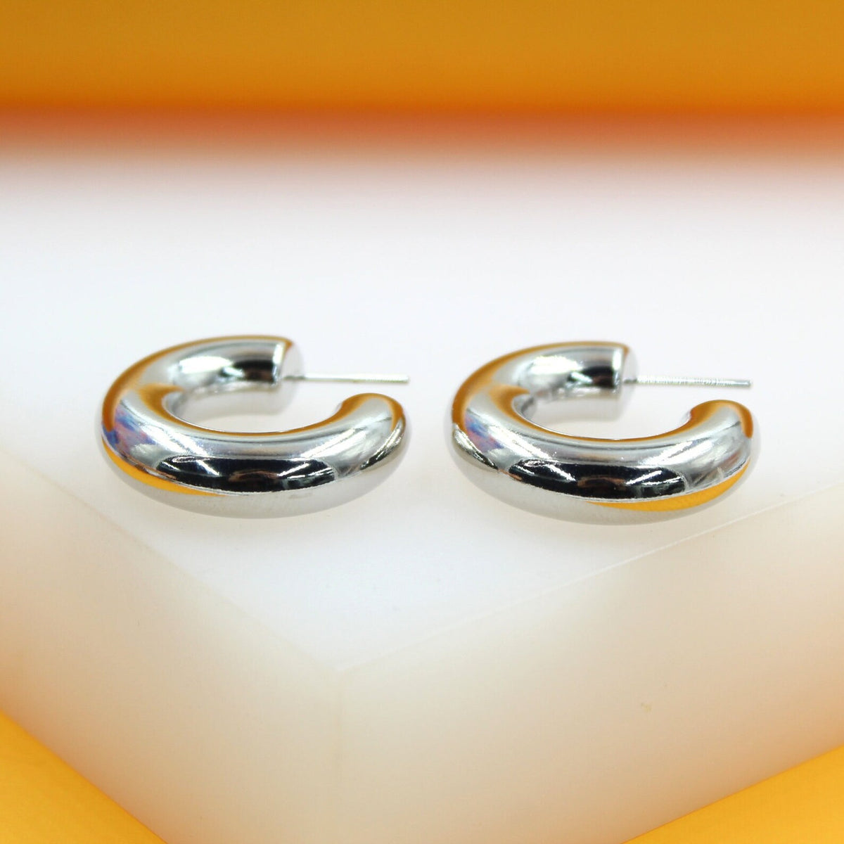 A pair of thick, polished silver hoop earrings rests on a white display block against an orange backdrop.