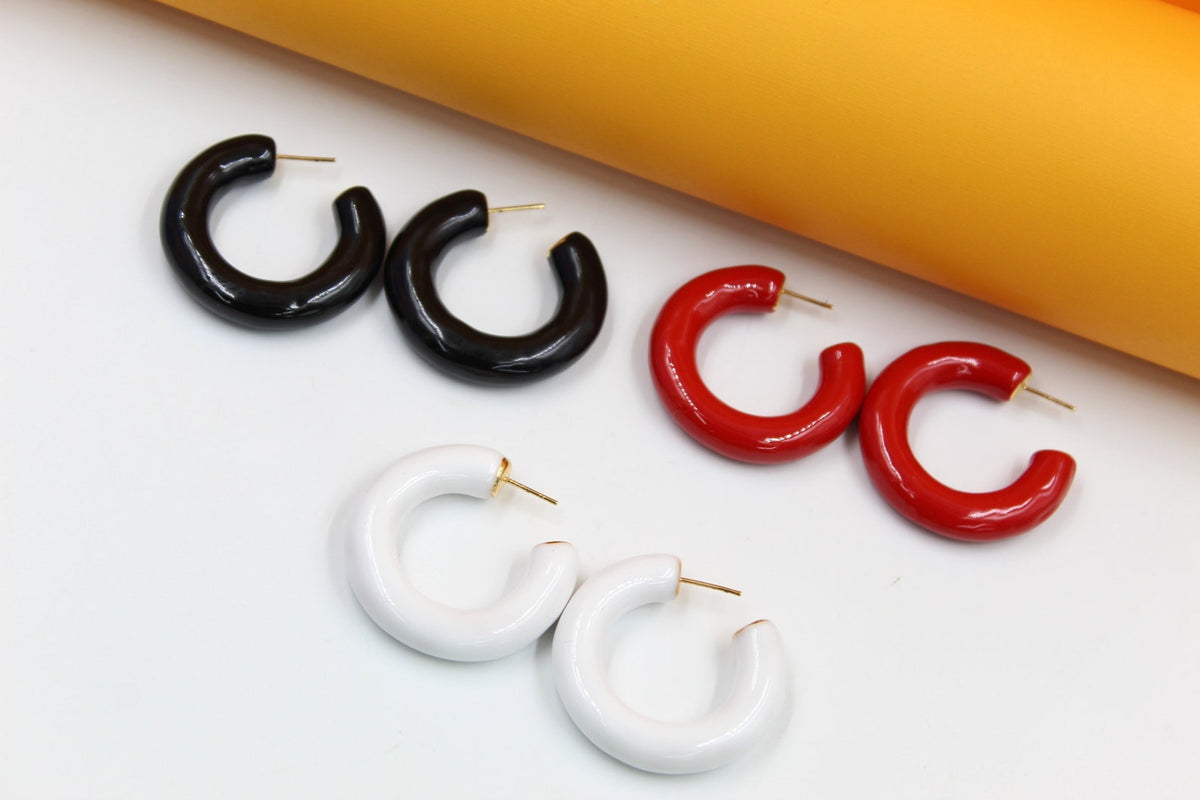 Three pairs of glossy, thick hoop earrings in black, red, and white rest on a white surface against an orange backdrop.