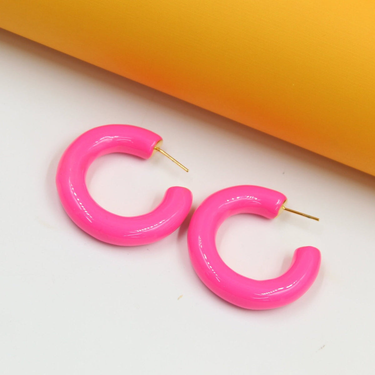 A pair of vibrant neon pink enamel hoop earrings rests on a white and yellow surface.