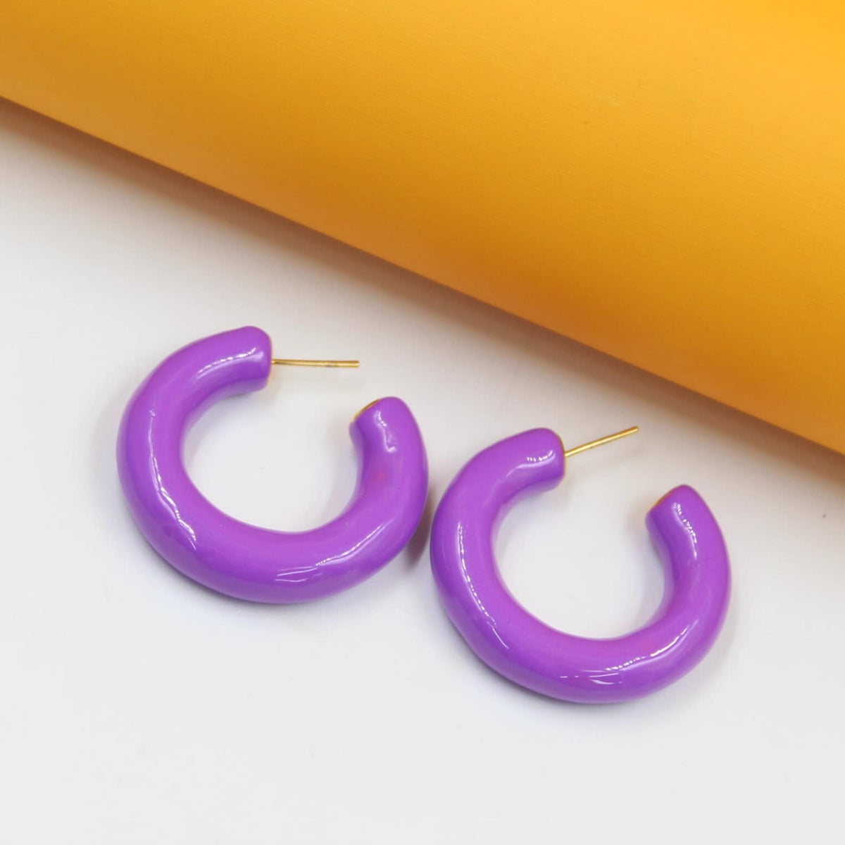 Glossy purple hoop earrings rest against a contrasting white and yellow background.