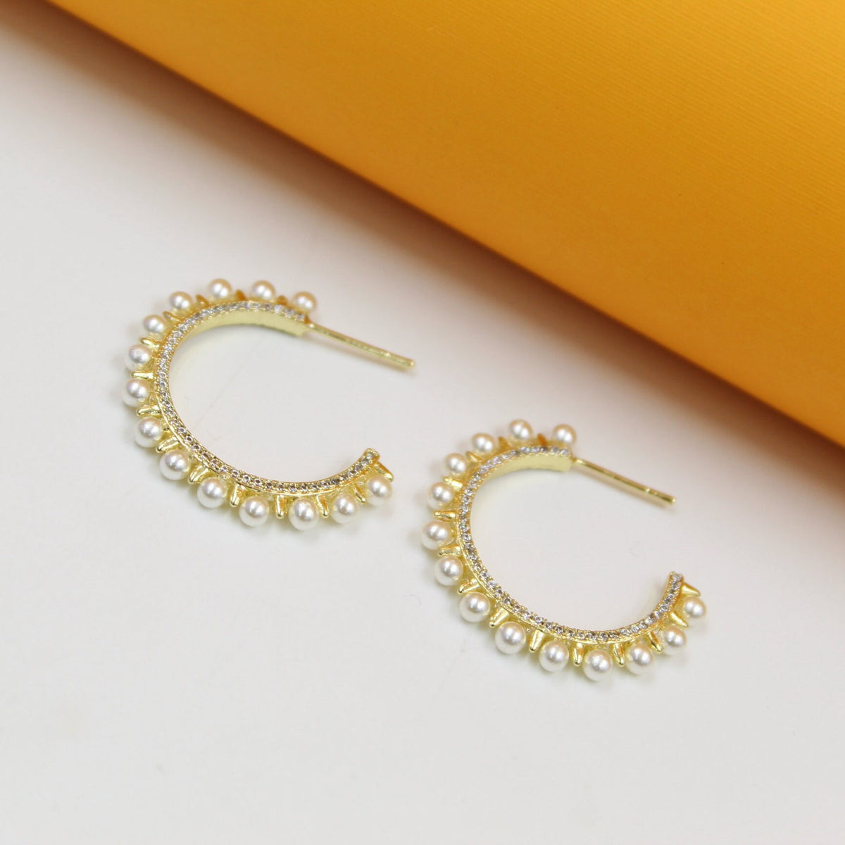 A pair of elegant gold hoop earrings adorned with shimmering pearls and sparkling crystals rests on a white and yellow surface.