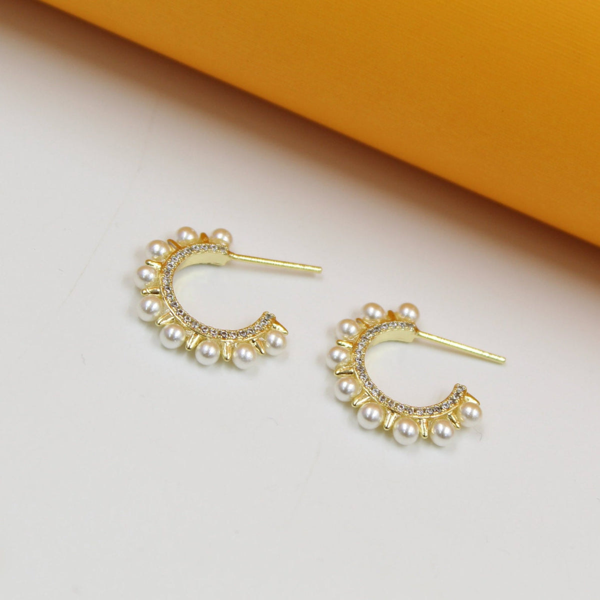 A pair of gold huggie hoop earrings features sparkling cubic zirconia and delicate white pearl accents against a neutral white and yellow background.