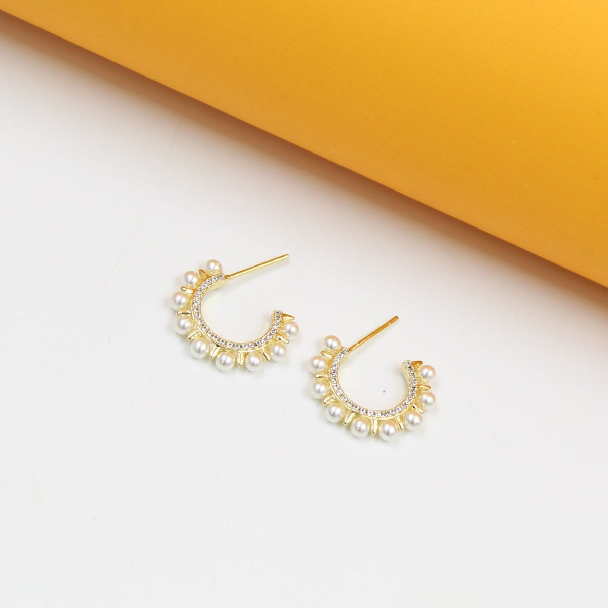 A pair of gold huggie hoop earrings decorated with delicate pearls and sparkling pave crystals rest against a white and yellow background.