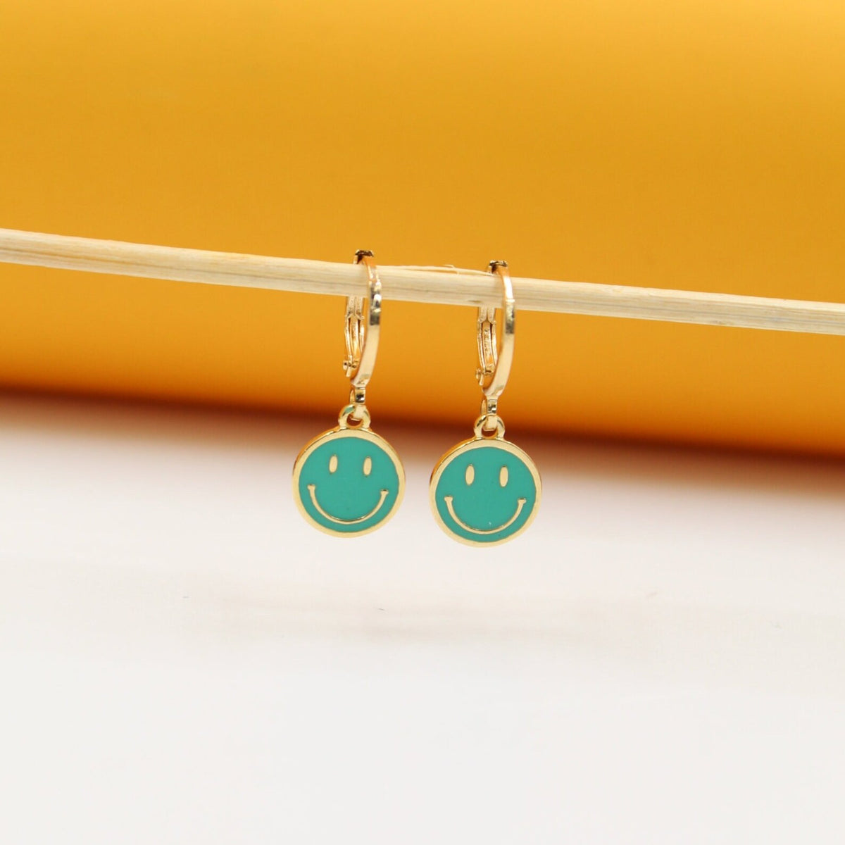 A pair of gold huggie hoop earrings featuring teal enamel smiley face charms hangs from a thin wooden stick against a warm yellow and white background.
