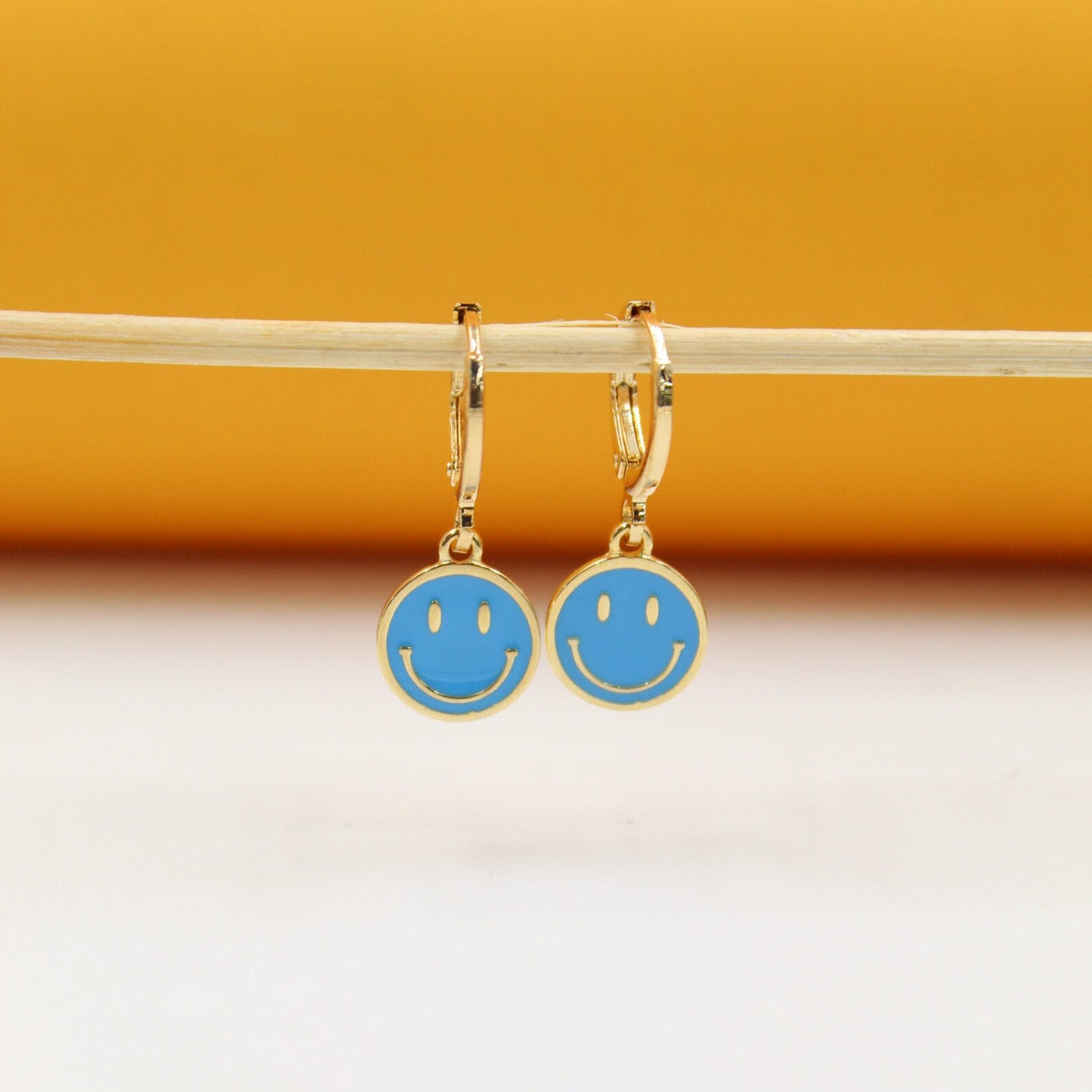 A pair of gold huggie hoop earrings with blue enamel smiley face charms hang from a wooden stick against a yellow and white background.