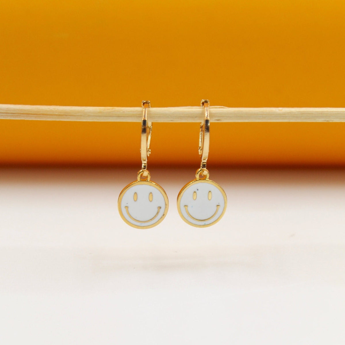 A pair of gold huggie hoop earrings featuring white enamel smiley face charms hang from a wooden stick against a warm orange backdrop.