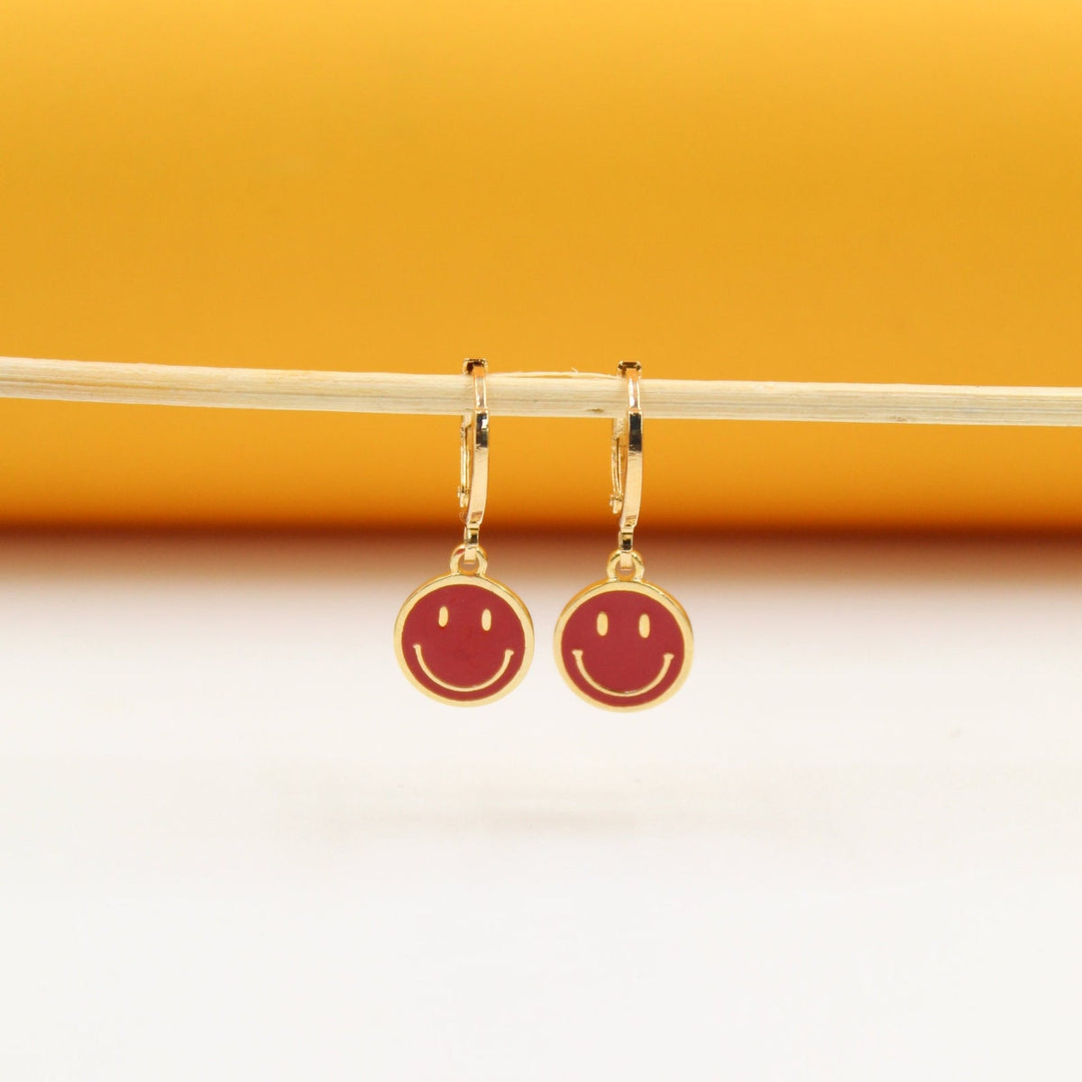 A pair of gold huggie hoop earrings featuring cheerful red smiley face charms hangs neatly from a thin wooden stick against a warm yellow and white background.