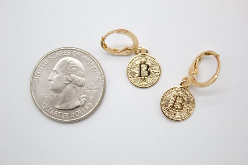 Two gold Bitcoin hoop earrings lie next to a US quarter on a white surface for size comparison.