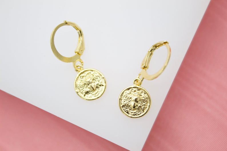 A pair of gold coin hoop earrings featuring an embossed Medusa motif rest on a white surface against a pink background.