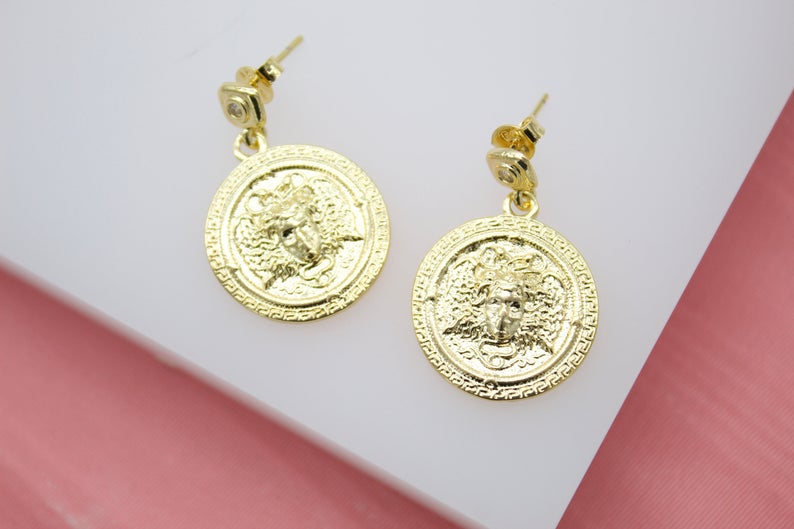 A pair of ornate gold Medusa coin dangle earrings rests on a white display surface.