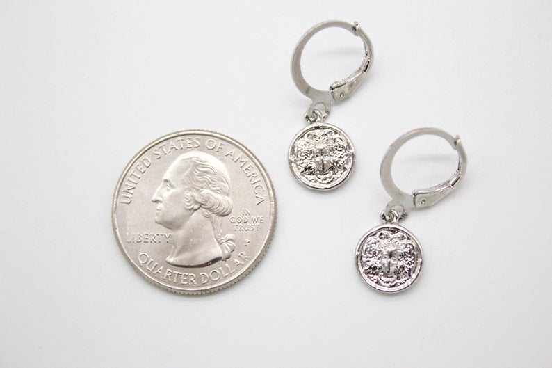 A pair of silver Medusa coin charm hoop earrings rests next to a US quarter coin for scale on a white surface.
