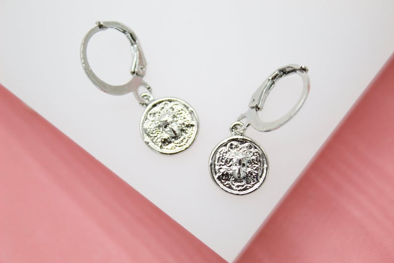 A pair of silver leverback earrings features dangling Medusa coin charms against a white and pink backdrop.