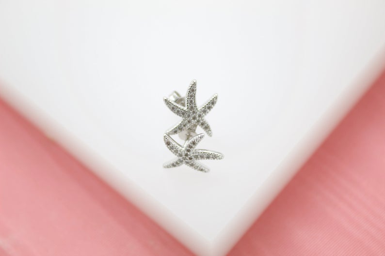 A sparkly silver starfish earring rests delicately on a white display against a soft pink background.