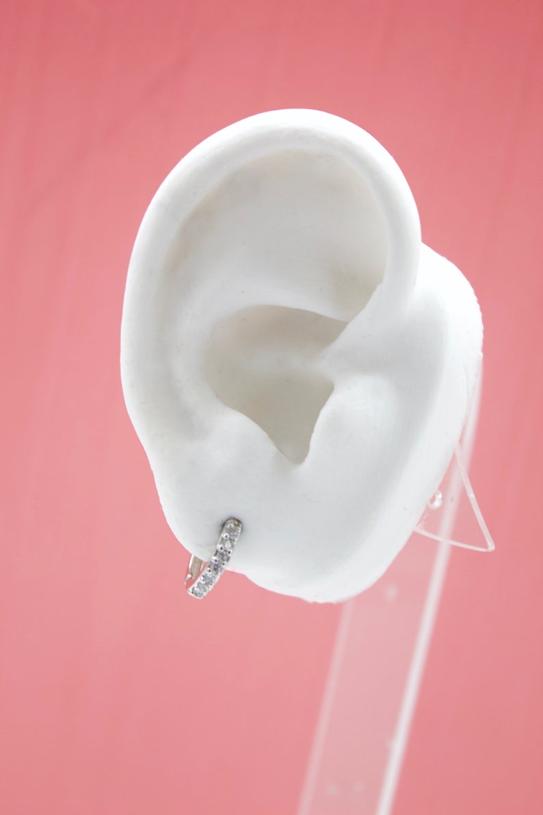 A sparkling diamond huggie hoop earring adorns the lobe of a white mannequin ear against a solid pink backdrop.