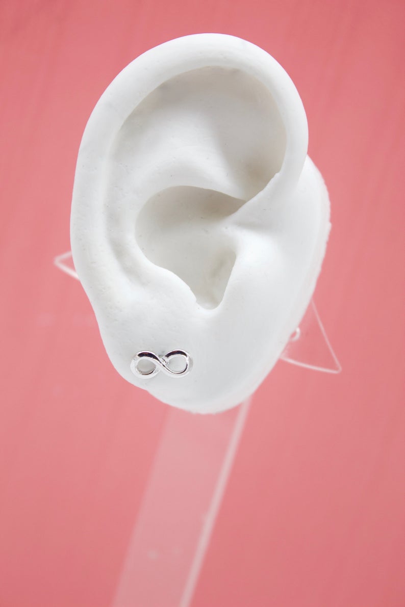 A silver infinity symbol stud earring adorns a white mannequin ear against a pink background.