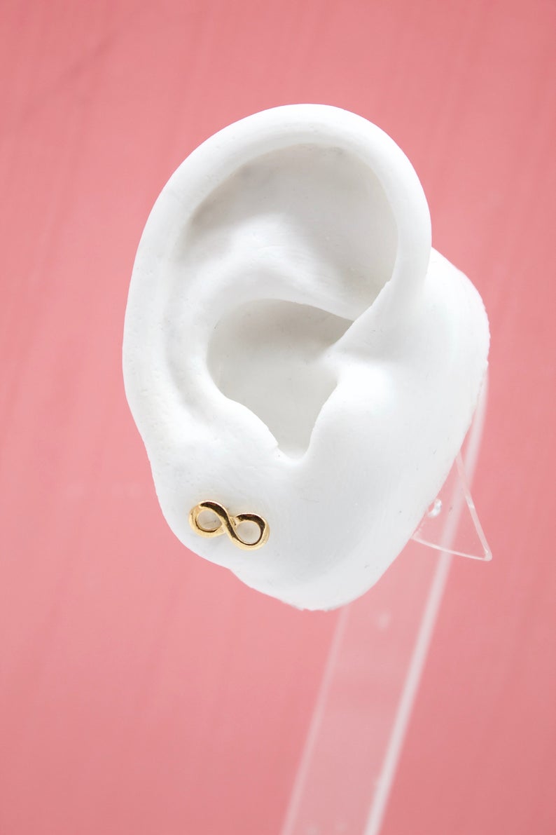 A white mannequin ear displays a delicate gold infinity sign stud earring against a pink background.