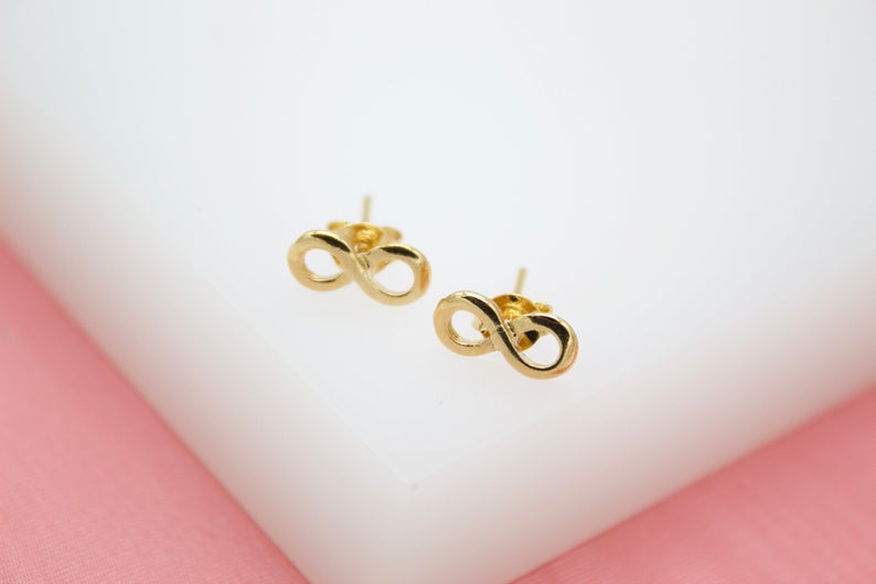 A pair of polished gold infinity symbol stud earrings rest on a white display surface.