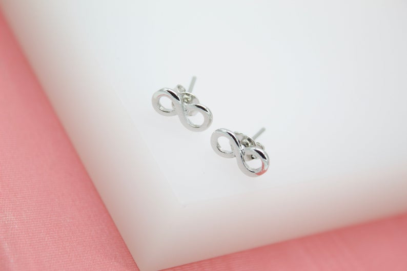 A pair of shiny silver infinity symbol stud earrings rests on a white display block against a soft pink background.