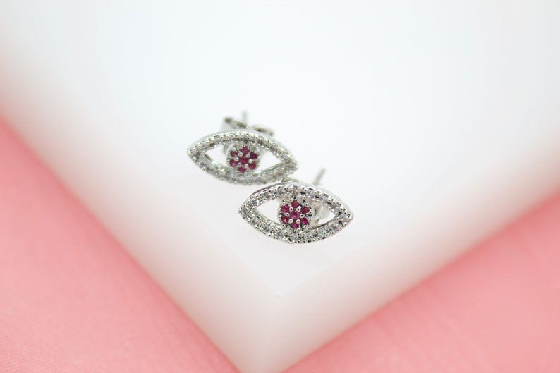 Two silver evil eye stud earrings sparkle with white crystals and pink center stones against a soft pink background.