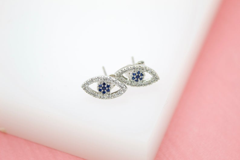 A pair of silver evil eye stud earrings featuring sparkling clear pavé diamonds and a central cluster of dark blue sapphires rests on a white display surface.