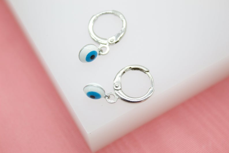 Two silver huggie hoop earrings dangle charmingly, each featuring a delicate evil eye charm with blue and white enamel.