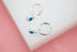 Two silver huggie hoop earrings dangle charmingly, each featuring a delicate evil eye charm with blue and white enamel.