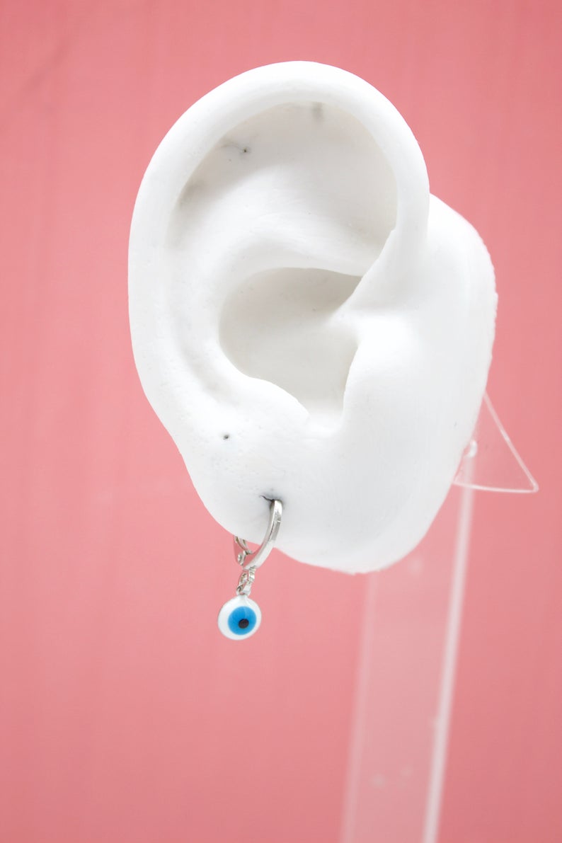 A white model ear displays a silver hoop earring featuring a blue and white evil eye charm against a pink background.