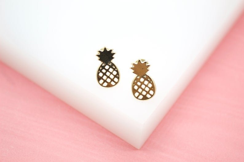 A pair of delicate gold pineapple stud earrings rests on a white display against a pink background.