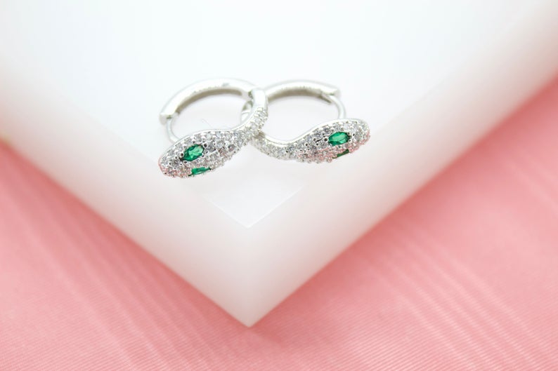A pair of silver snake hoop earrings encrusted with clear cubic zirconia stones and featuring vibrant green emerald eyes rests on a white display block.