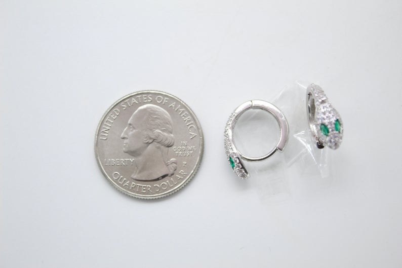 A pair of silver snake hoop earrings with crystal pavé and green gem eyes rests next to a US quarter coin for scale.