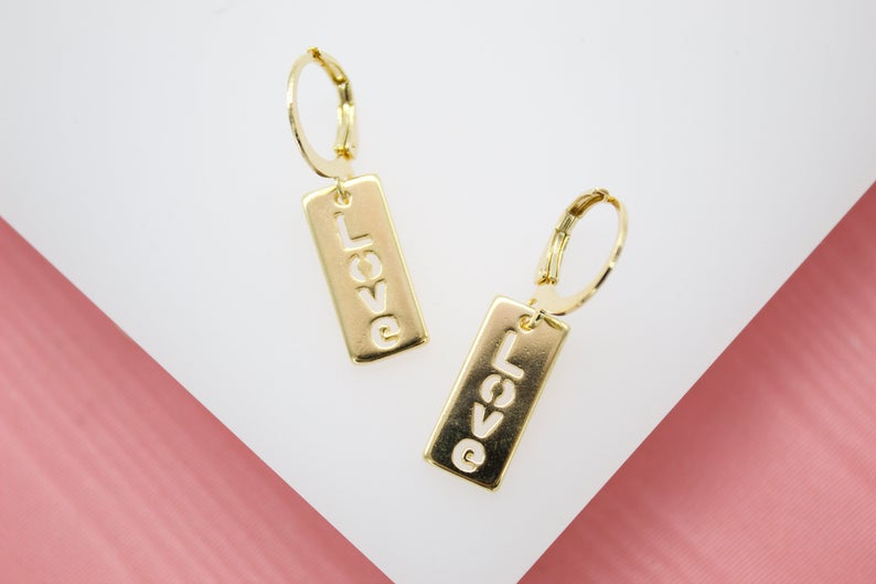A pair of gold dangle hoop earrings featuring rectangular charms with the word "LOVE" cut out lies on a white and pink surface.