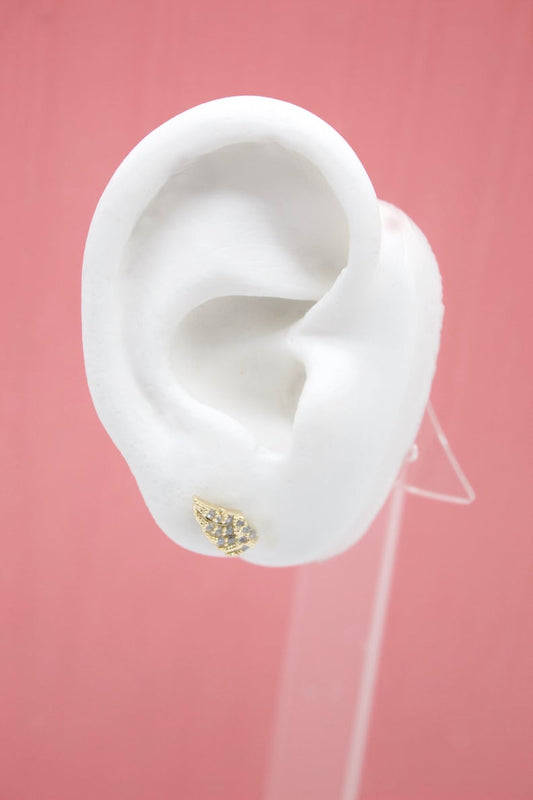 A white mannequin ear on a pink background displays a small gold wing stud earring encrusted with tiny crystals.
