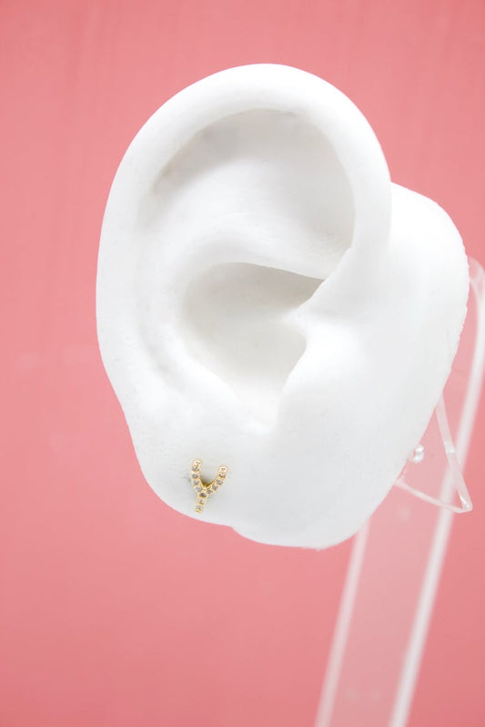 A white mannequin ear displays a sparkling gold initial Y stud earring against a pink background.