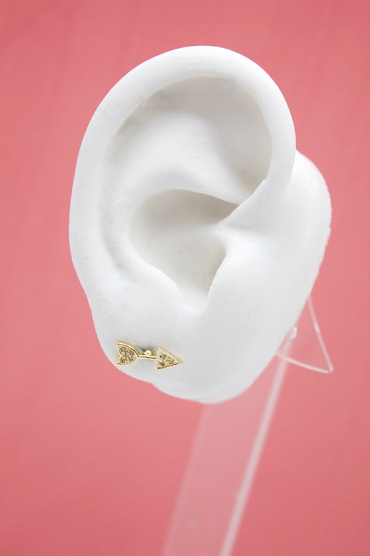 A white model ear showcases a gold arrow stud earring embellished with small sparkling stones against a solid pink background.