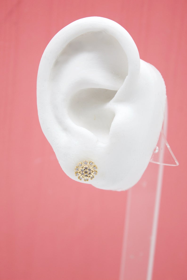 A white model ear on a pink background displays a sparkling gold flower stud earring encrusted with tiny clear crystals.