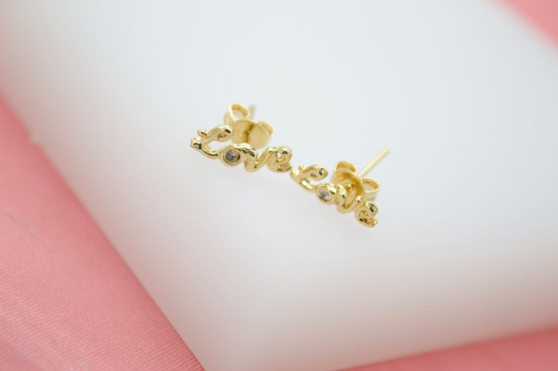 A pair of gold script love earrings sparkle on a white display surface.