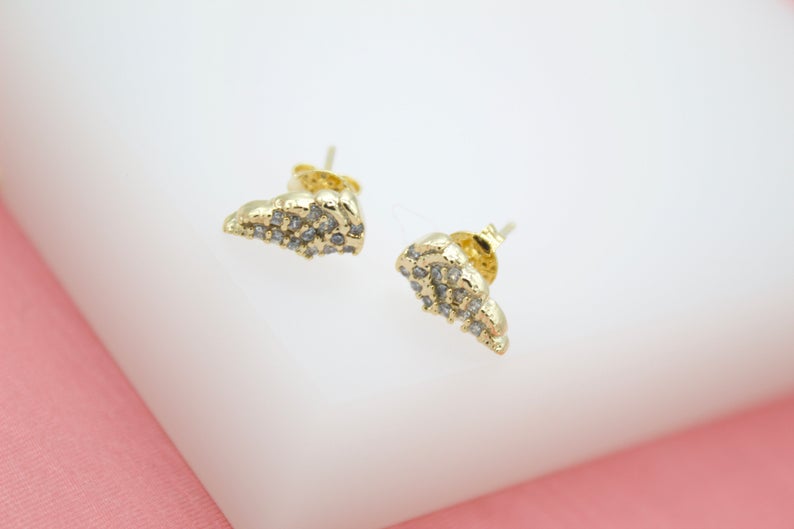 A pair of gold croissant stud earrings sparkle against a soft pink and white background.