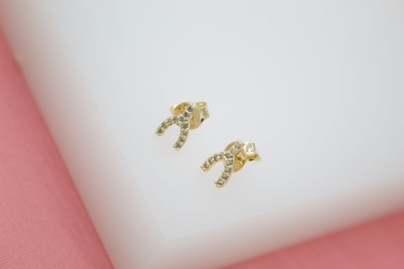 A pair of gold wishbone stud earrings encrusted with sparkling cubic zirconia stones rests on a white display block.