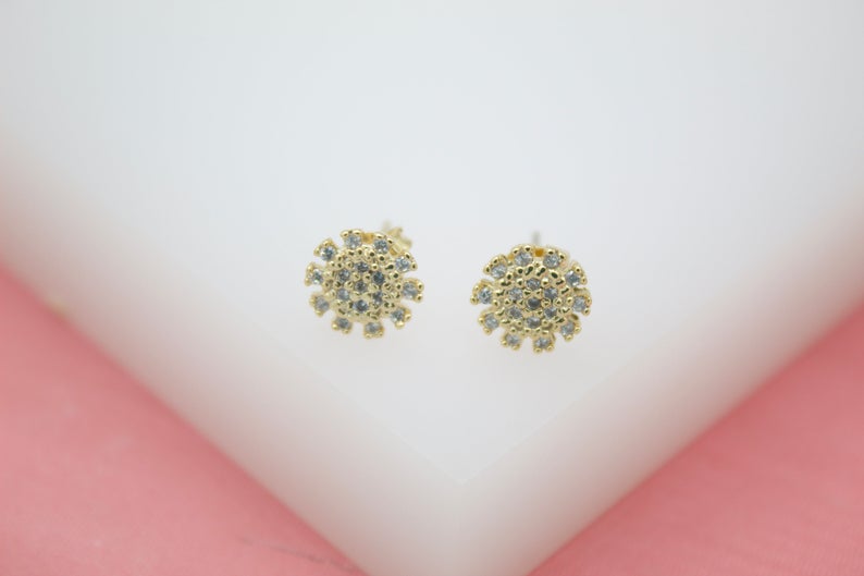 A pair of sparkling gold sunburst stud earrings encrusted with tiny cubic zirconia stones rests on a white display surface.
