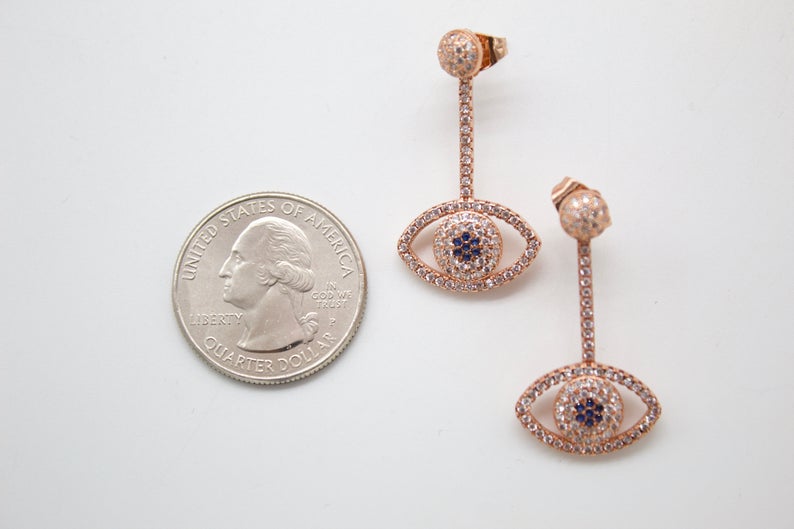 A pair of rose gold evil eye drop earrings embellished with sparkling cubic zirconia rests next to a US quarter coin for scale.