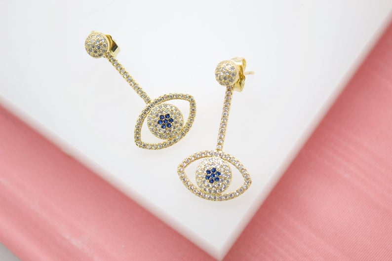 A pair of gold evil eye drop earrings embellished with sparkling white cubic zirconia stones and central blue crystals rests on a white display surface against a pink background.