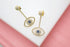 A pair of gold evil eye drop earrings embellished with sparkling white cubic zirconia stones and central blue crystals rests on a white display surface against a pink background.