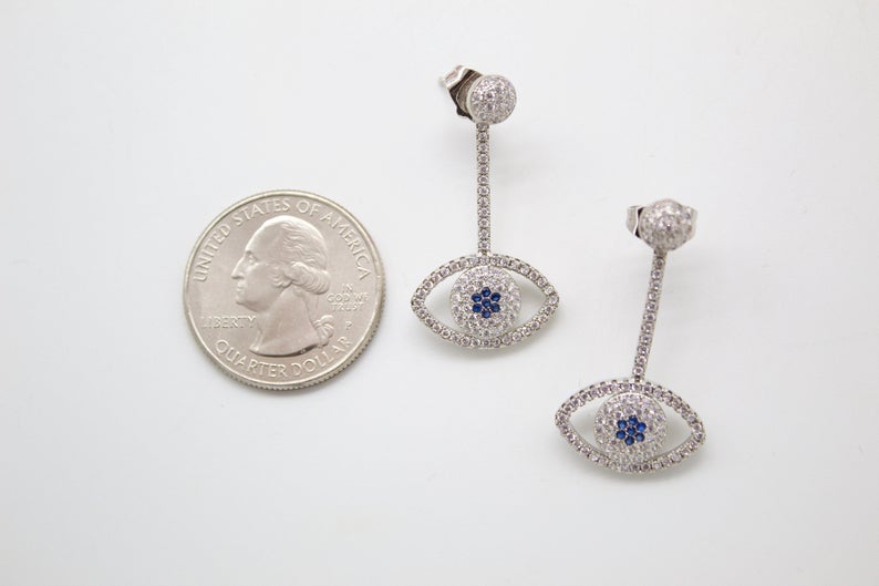 A pair of sterling silver evil eye drop earrings embellished with pave-set cubic zirconia and blue sapphire stones rests next to a US quarter coin for scale.