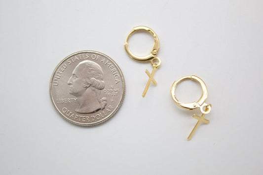 A pair of gold cross hoop earrings rests next to a US quarter coin for scale on a light gray surface.