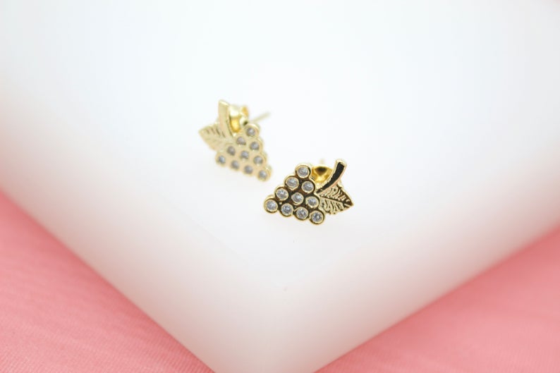 A pair of gold grape cluster stud earrings sparkle with embedded cubic zirconia stones on a white and pink surface.