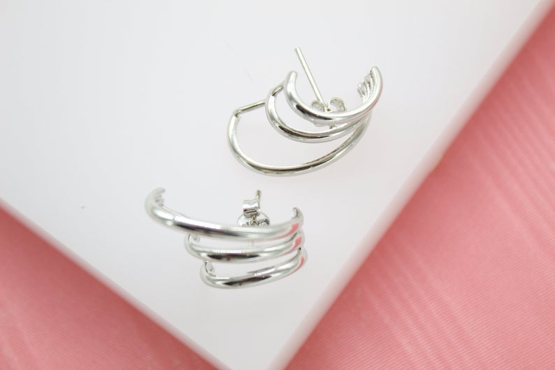 A pair of multi-hoop silver earrings rests on a white display surface against a pink background.