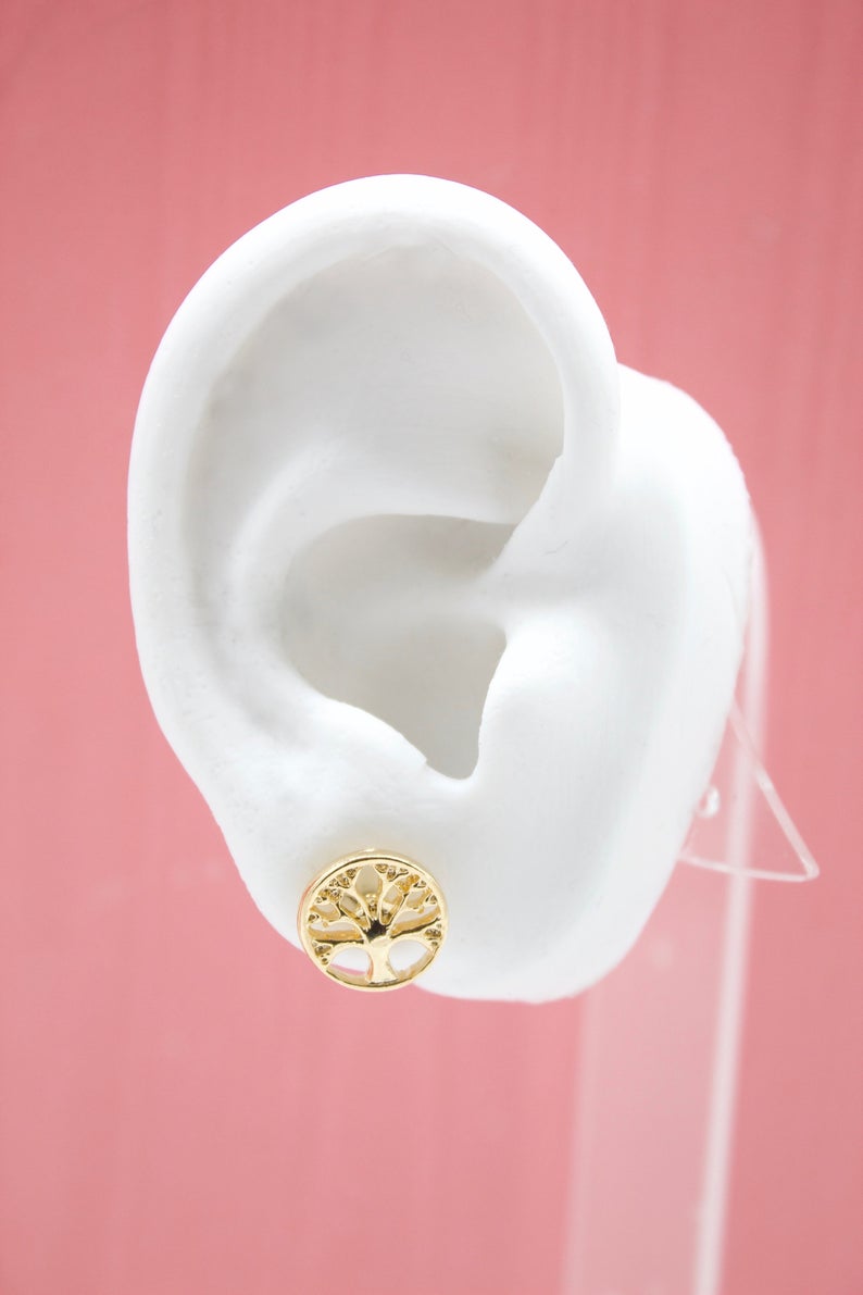 A white mannequin ear displays a round gold tree-of-life stud earring against a pink background.
