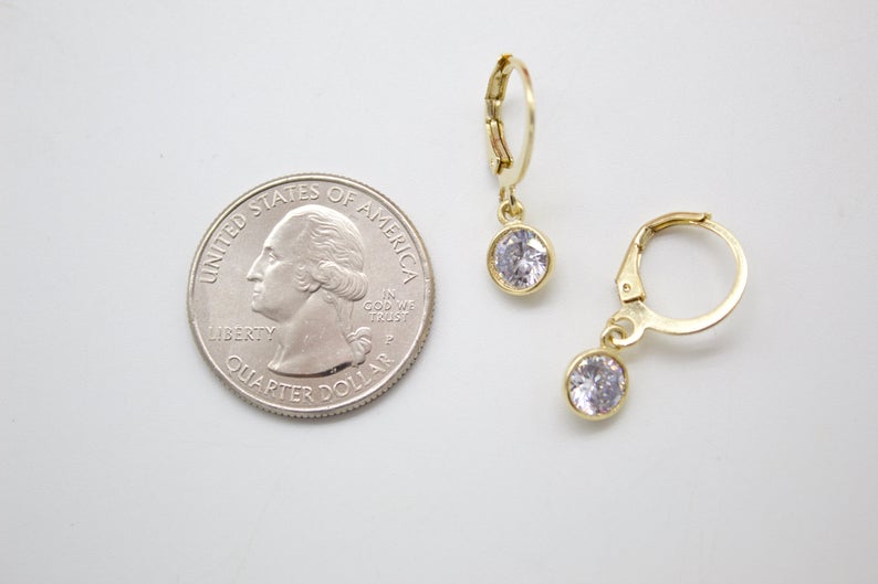 A pair of gold leverback earrings with dangling cubic zirconia stones rests next to a US quarter coin for scale on a white surface.