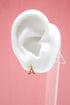 A white mannequin ear displays a gold Eiffel Tower stud earring with pink enamel detail against a solid pink backdrop.